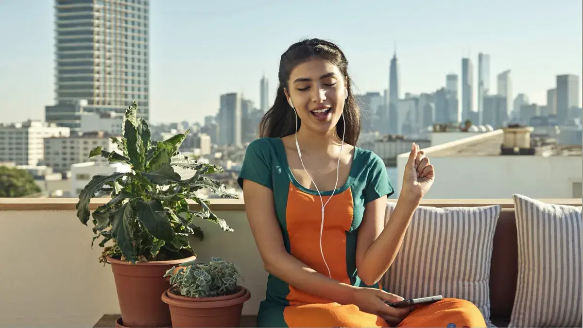 A young Indian woman on a sunny terrace with earphones, practicing spoken English alone at home by speaking out loud